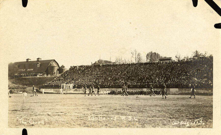 A crowded audience watches the football game between the University of Idaho and Washington State College on the field next to the cattle barn.