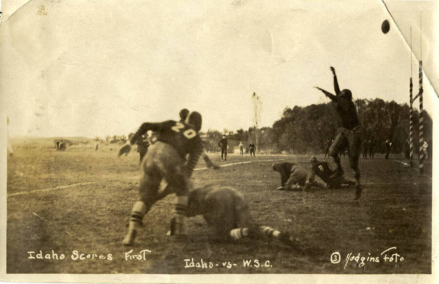 An action shot taken during a football game between the University of Idaho and Washington State College captioned with, 'Idaho Scores First.' The photo captured a man jumping for the ball while other players are on the ground recovering from tackles.