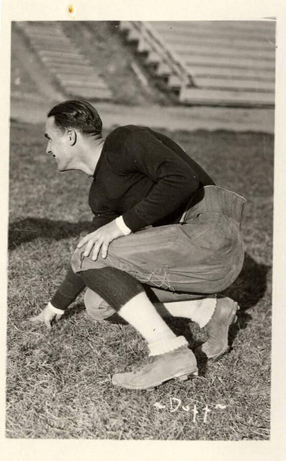 A football player crouches on the field in front of the bleachers in a classic uniform of the time with cleats, legwarmers, pants with a thick waist band and lacing up the back and a black shirt. The photograph is captioned with '~Dutt~' which could be referencing the player or the photographer