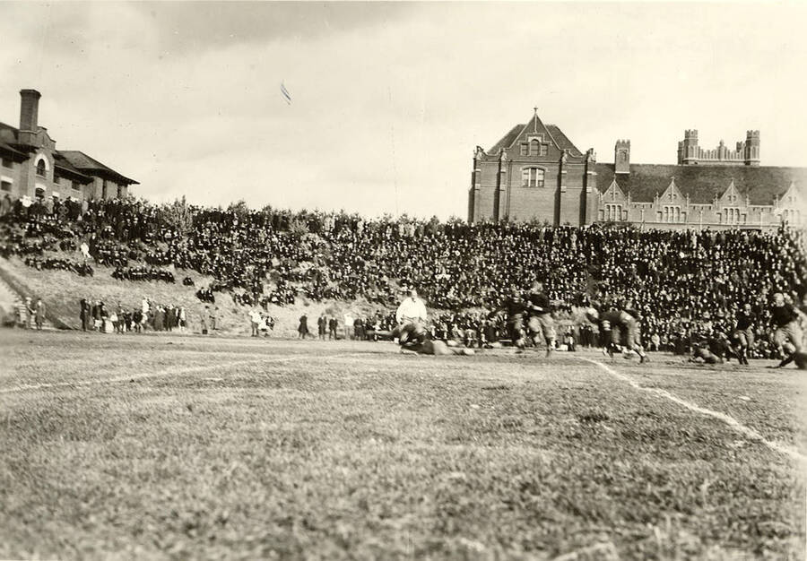 The audience, situated on the bleachers, hill, and sidelines, watch the football game as the referee and players rush to a tackle.