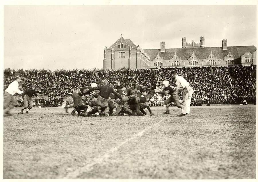Referees monitor as players rush in to turn a tackle into a dogpile while the crowded audience watches from the bleachers.