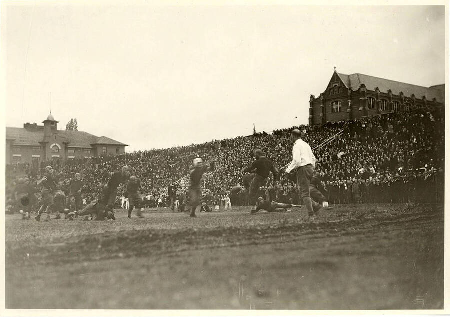 Football players on the field watch expectantly as the kicker kicks the ball and the opposing team tries to interfere. The audience is packed along the side and you can see peaks of surrounding buildings.