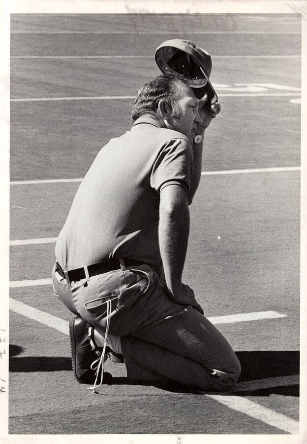 A University of Idaho coach kneels on the sideline with a shoelace hanging out of his back pocket, watch twisted onto the inside of his wrist, and hat tilted towards the football field.