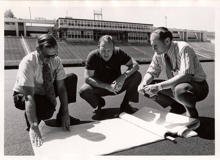 Three men, including a University of Idaho football coach, crouch on the football field while analyzing a large set of plans.