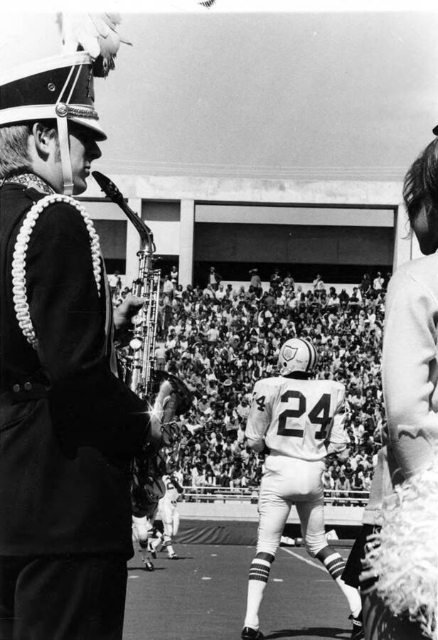 A photograph peaking through the bodies of a saxophone player and a cheerleader to view a football player and crowded stands.