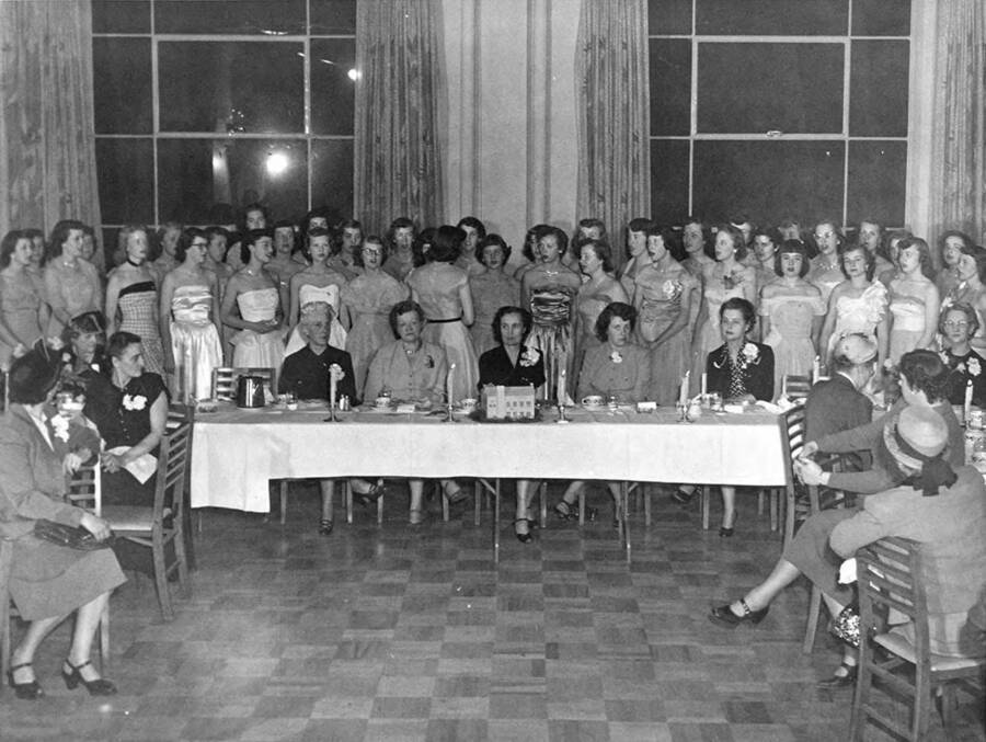 Formally dressed women stand and sit around a table at the Kappa Alpha Theta house during the Kappa Alpha Theta reunion.