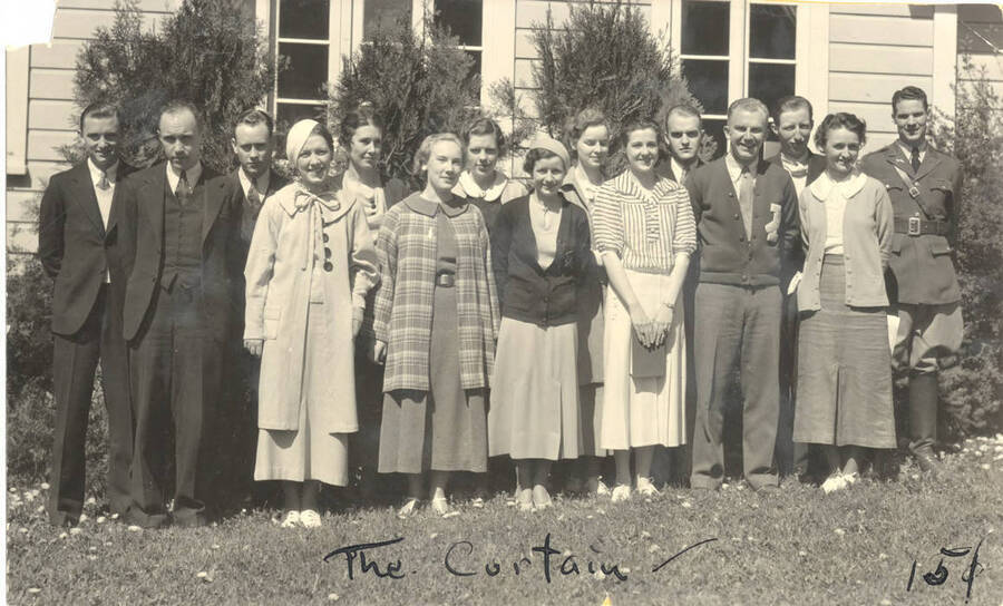 The Curtain members pose for a photo together next to a building. The Curtain Club is the dramatics honorary club at the University of Idaho. Individuals identified as listed. Back: Earl Bopp, Harrod Enking, Dorothy Ward, Rosanne Roark, Ethlyn O'Neal, Robert Herrick, Raphael Gibbs, Clayne Robison; front: Fred Blanchard (head of dramatics), Ruth Lyon, Marion Dresser, Margaret Moulton, Jean Ricker, Lloyd Ruitel, Nina Varian. Caption reads: 'The Curtain ~ 15'.