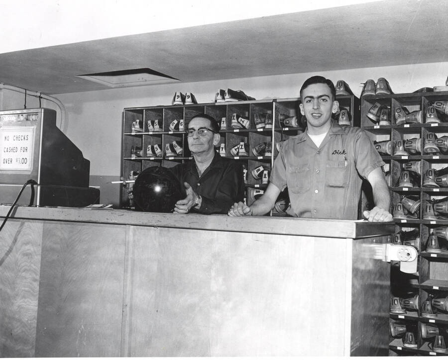 Student Union Building employees Jim Bowlby (game room manager) and Dick Smith (maintenance manager) standing behind the bowling checkout counter. Jim Bowlby holds a bowling ball.