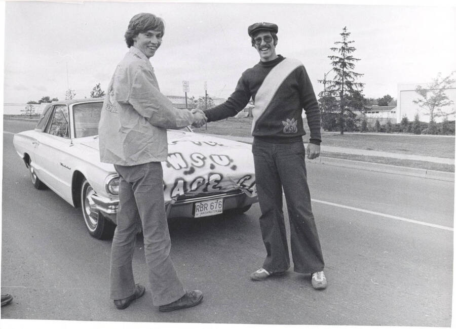 Two unidentified individuals shake hands in front of a Thunderbird bearing a banner that declares it the 'U of I WSU Pace Car'.