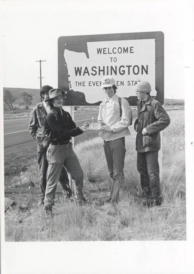 Four men hold a license plate in front of the 'Welcome to Washington' sign.