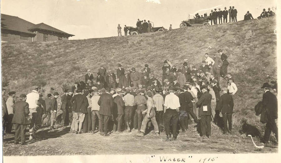 A group of male students gather around a crowd that is blocked from view. Spectators watch from the crest of the hill above the gathering. Caption reads: ''Water' 1915.'