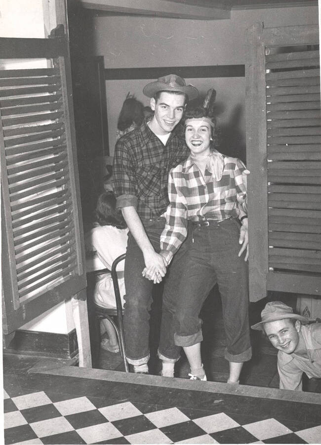 An unidentified greek couple dressed in Western themed clothing poses by a saloon-style door while another member peeks out from beneath the door.