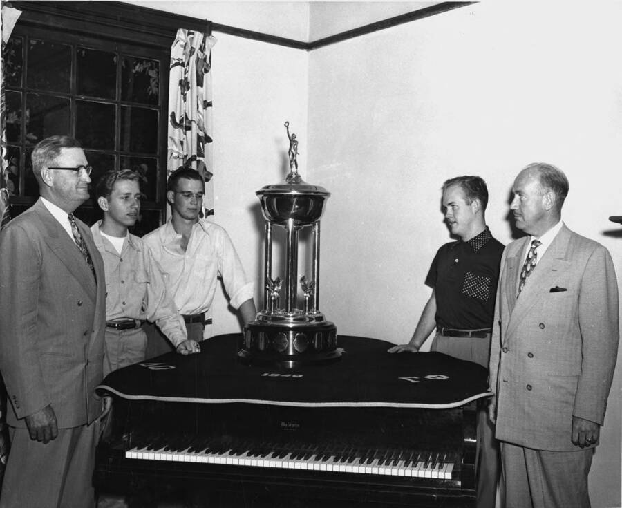 A group of men stand around the Sigma Chi scholarship trophy, which is on top of a piano.Pictured left to right: President Jesse Buchanan, Stanley Swanson, William Mahlik, Richard Coulter, Carl P. Clare