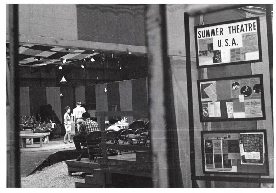 Theatre students rehearse inside an auditorium. Newpaper clippings are framed on a wall with one of them reading: 'Summer Theatre U.S.A.'