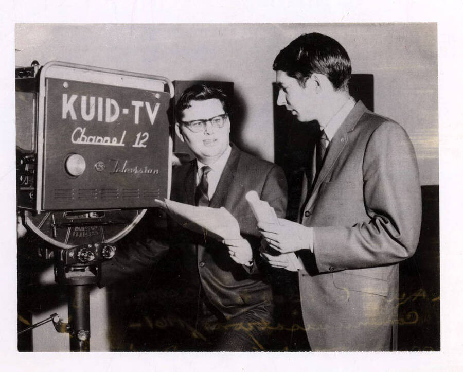 Dr. Gordon Law and Peter Haggart stand next to a video camera that reads: 'KUID-TV Channel 12', holding papers and talking to each other.