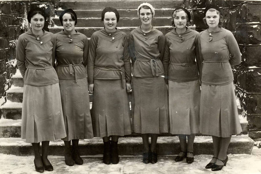 Vandalettes pose for a group photograph wearing long coats at the base of the Women's Gymnasium staircase during winter. Individuals identified from left to right: Laura Brigham, Ruth Brody, Elizabeth Thompson, Edna Scott, harriet Baken and Bernice Smith.