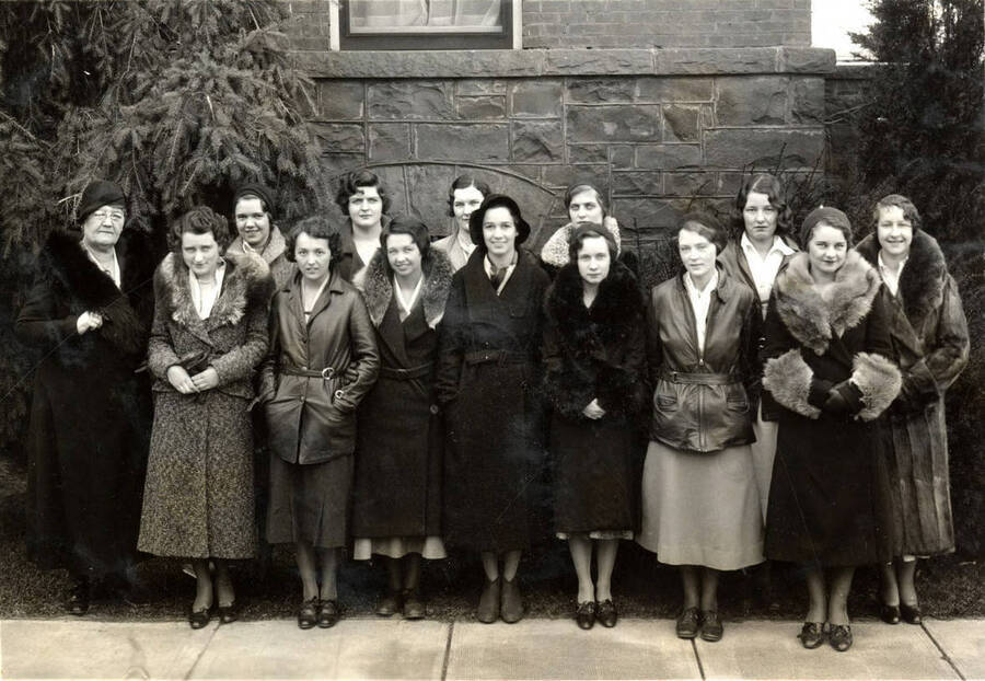 The Executive Board of Associated Women Students pose for a photo together wearing long coats in front of the Women's Gymnasium. Individuals identified as listed. Permeal French (Adv.), Teresa Connaughton, Elsa Eisinger, Irene Luke, Catherine O'Brien, Dorothy Lindsey, Marion Fry, Maude Galloway, Pearl Walters, Margaret Scott, Ruth Cook and Mae Belle Donaldson