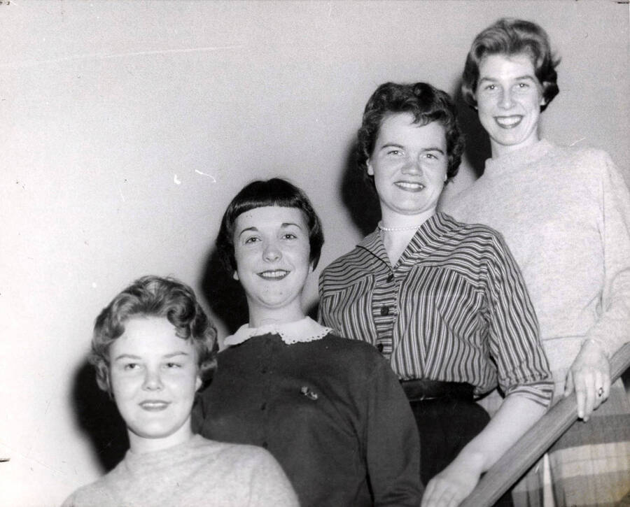 The four officers of Associated Women Students pose for a photo together on the staircase of an unidentified building. Individuals identified as listed. Deanna M Geersten (vice president), Nancy R. Campbell (treasurer), Kay Zenier (president) and Irene Scott (secretary).