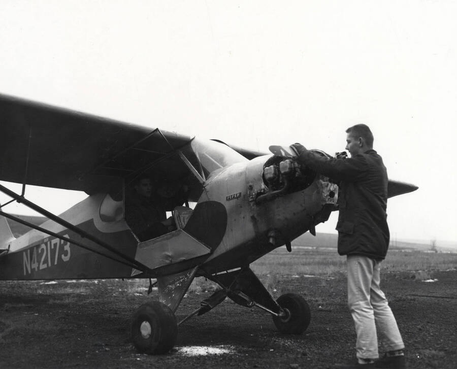 A member of the Vandal Flying Club starts the propeller on a Piper aircraft while another member sits in the cockpit.