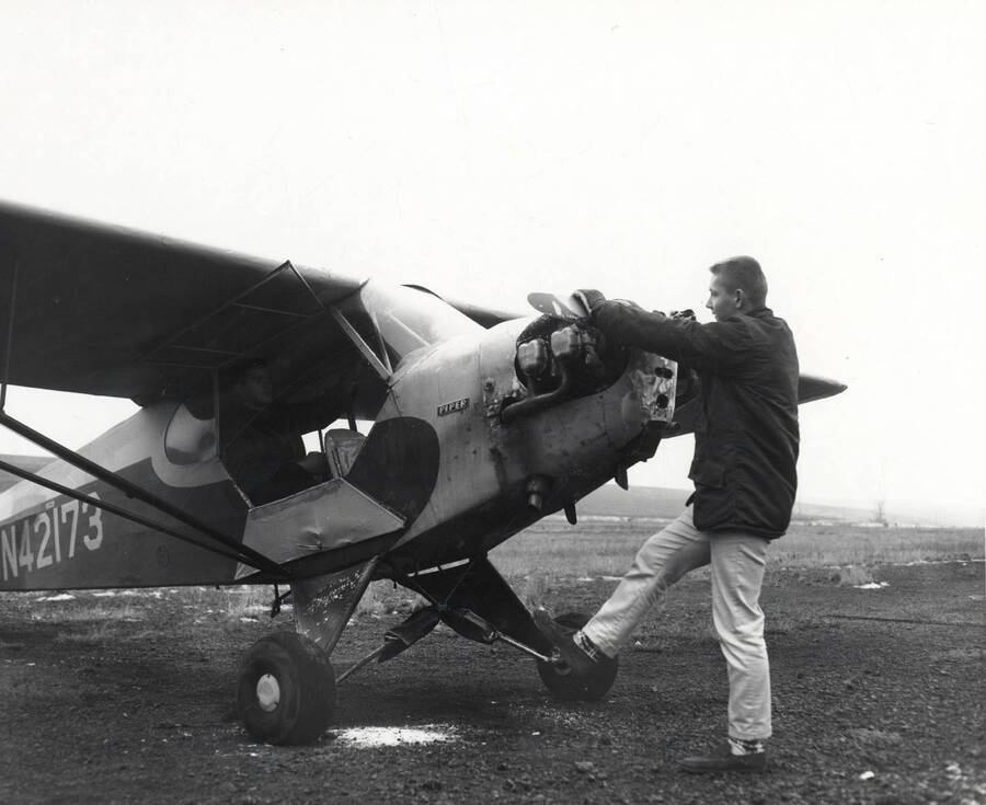 A member of the Vandal Flying Club starts the propeller on a Piper aircraft while another member sits in the cockpit.