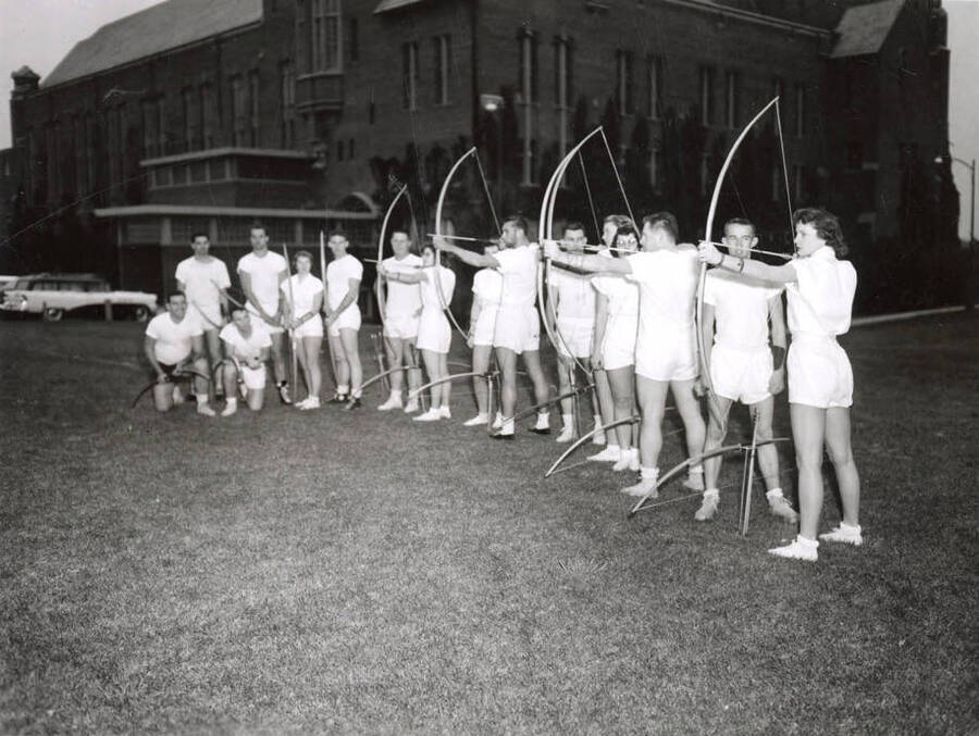 A group of students line up behind the Memorial Gymnasium during archery class. Five students practice their draw and release while the rest of the students watch.