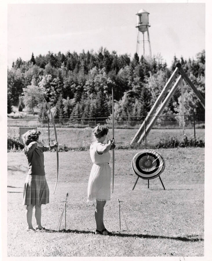 Two women aim at targets with drawn bows. The 'I' tower is visible in the background.