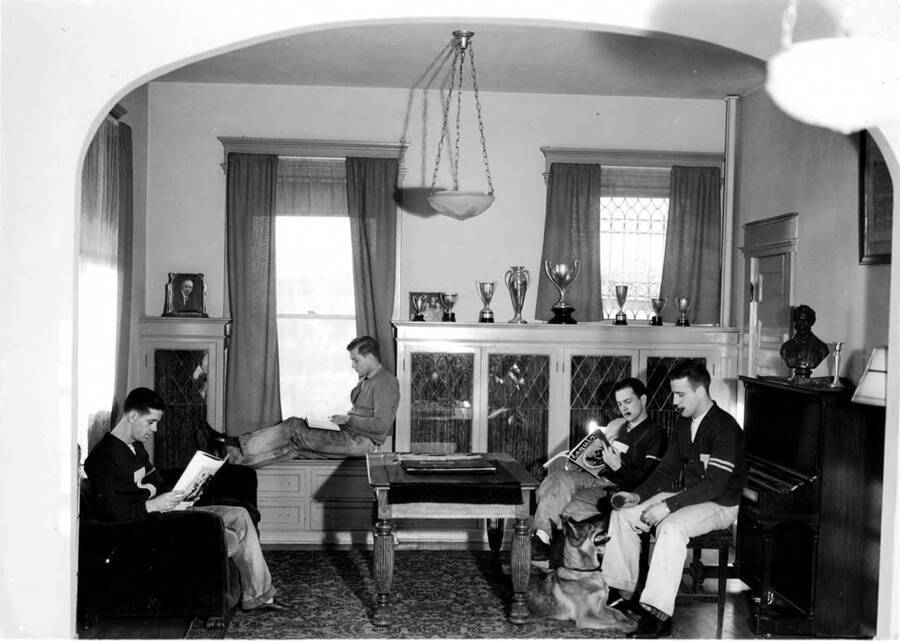 Four men sit in the living room of Tau Kappa Epsilon house at 616 S. Jefferson. Three men are reading and one tends to a dog.