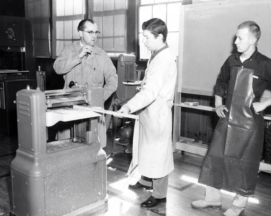 A professor instructs Industrial Arts Education students on using the electric planer while one student holds a piece of wood up to the machine.