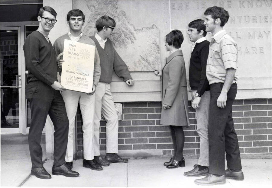 Gary Garnand, Fred Finlayson, Mike Hunter, Mary Kay Holden, Bill Flandro, and Lee McCollum stand outside the library and pointing towards Pocatello on the Idaho relief map during 'All Idaho Week.'
