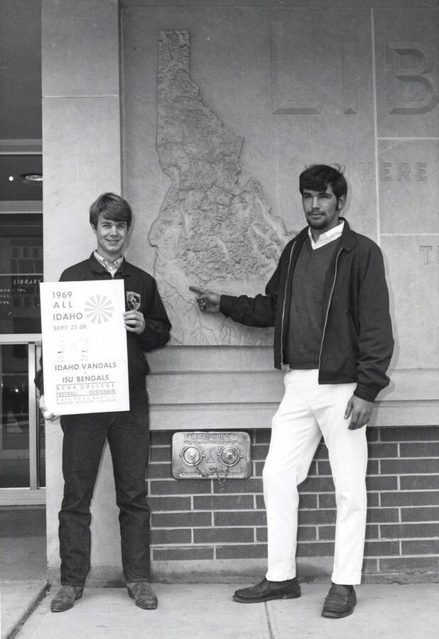 Two men hold a sign outside the library while pointing to the Boise area on a relief map of Idaho located on the outside of the library.