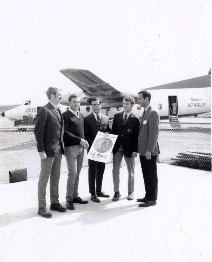 ASUI President Jim Willms, Scott McClure, Jim Mottern, Lee McCollum, and Senator John Mix stand outside of an airport runway with a sign for 'All Idaho Week.'