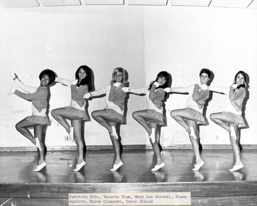 A group photograph of the Pom-Pom Girls lined up holding a cheer pose. Individuals identified from left to right: Patricia Kido, Valerie Pllum, Mary Lee Strobel, Diana Aguirre, Karen Clements, Carol Gibson.