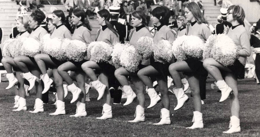 The Pom-Pom Girls perform a cheer on MacLean Field during a football game. The Vandal Marching Band performs in the background behind them.