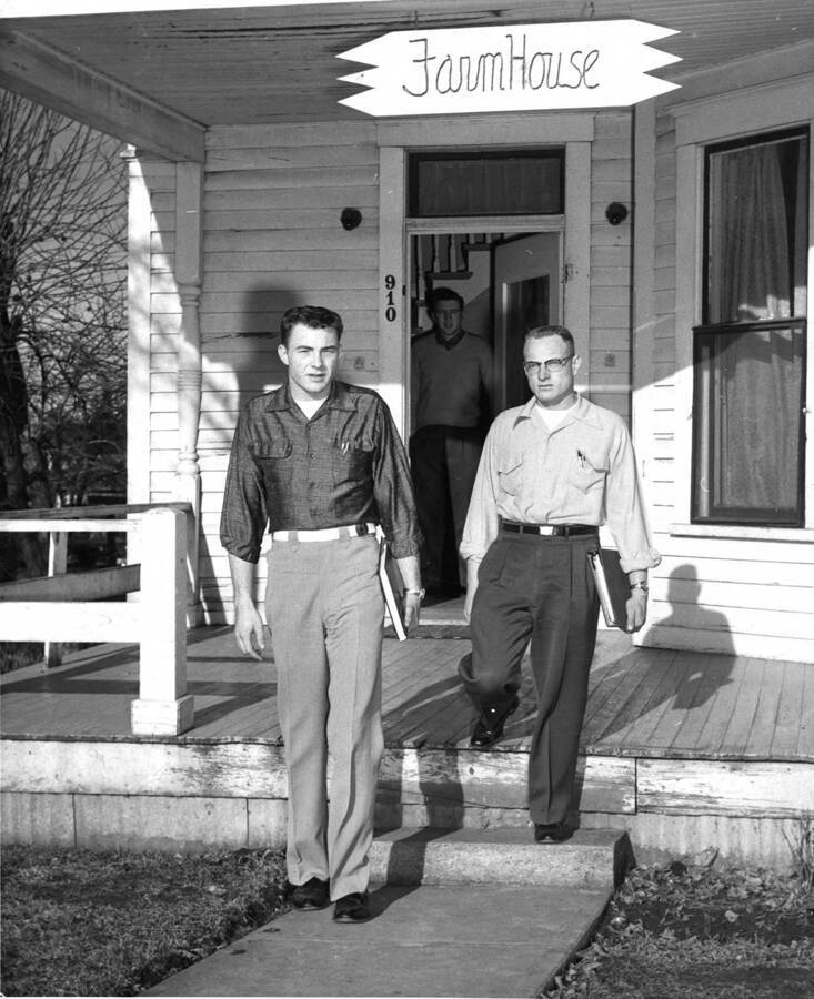Two men stand in front of the porch of Farmhouse fraternity while one man stands in the doorway.