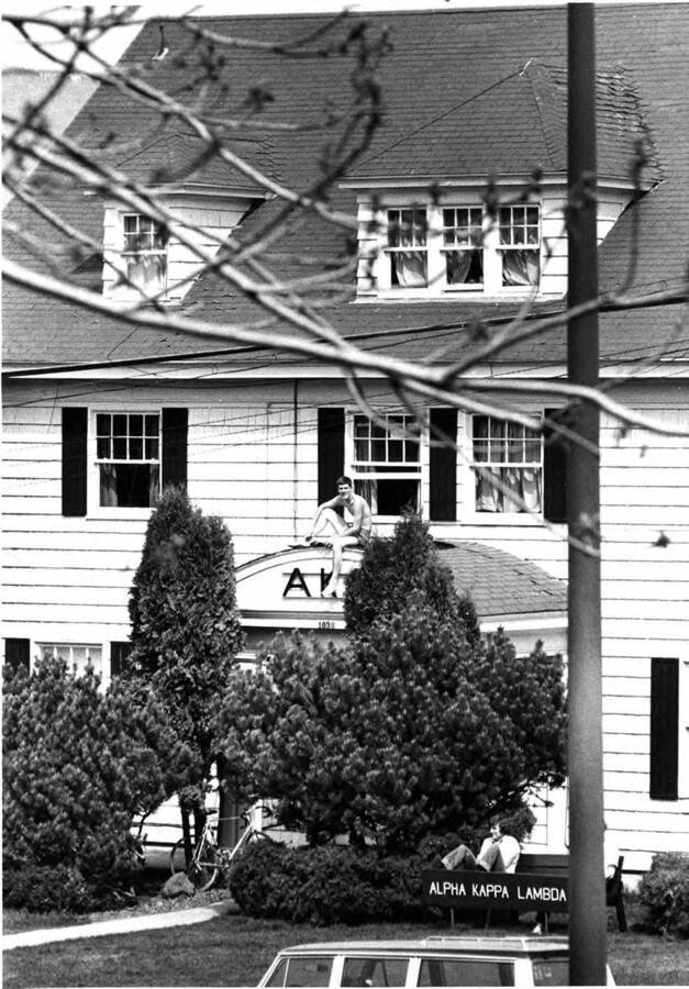 Photo of two men sitting outside Alpha Kappa Lambda; one sits over the entrance of the building, and one sits on a bench near the entrance. The house was previously used by Gamma Phi Beta (1917-1958) and Alpha Gamma Delta (1958-1969).