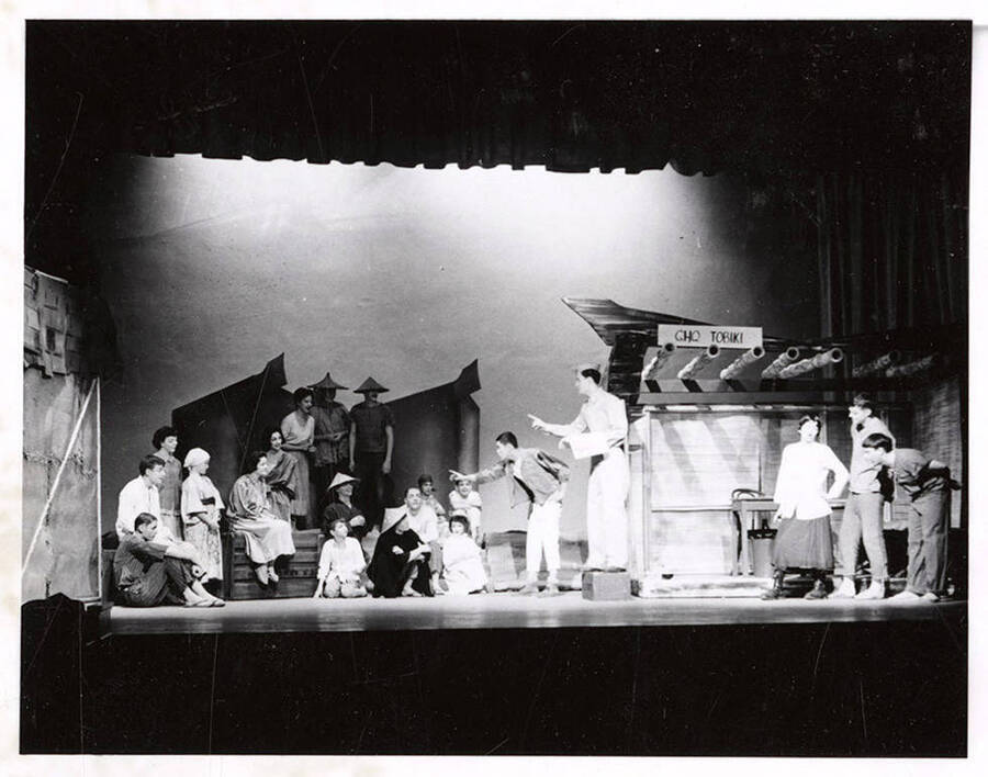 Students perform a group scene during a drama production of 'Teahouse of the August Moon.' The actors are costumed as Japanese villagers and American soldiers. Individual performers are identified on the photograph's mount. University of Idaho