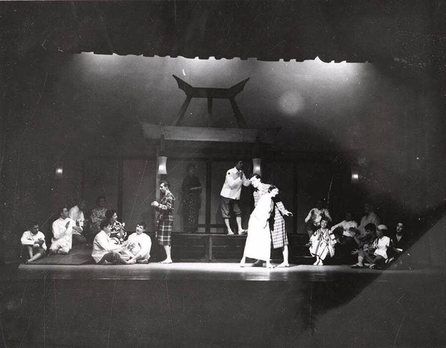 Students perform a group scene during a drama production of 'Teahouse of the August Moon.' Actors are costumed as Japanese villagers and American soldiers. Individual performers are identified on the photograph's mount. University of Idaho