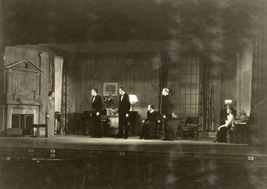 During the drama production of 'Dangerous Corner,' men and women stand on stage facing one woman. Cast: Robert Herrick as Gordon Whitehouse, Dorothy Pierce as Freda Chatfield, Eldred Stephenson as Robert Chatfield, Alline King as Olwen Peel, Paul Selby as Charles Stanton, Nina Varian as Betty Whitehouse, Ethlyn O'Neal as Maud Mockridge