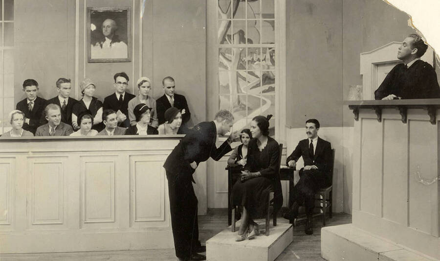 A man bows to a seated woman as others look on during the drama production of 'Ladies of the Jury.' Cast: Rosamond Tenney as Mrs. Crane, Edwin Ostroot as judge, Jack Blair and Casady Taylor as lawyers, Dorothy Menzies as defendant on trial, Naomi Randall as maid who testifies, etc.