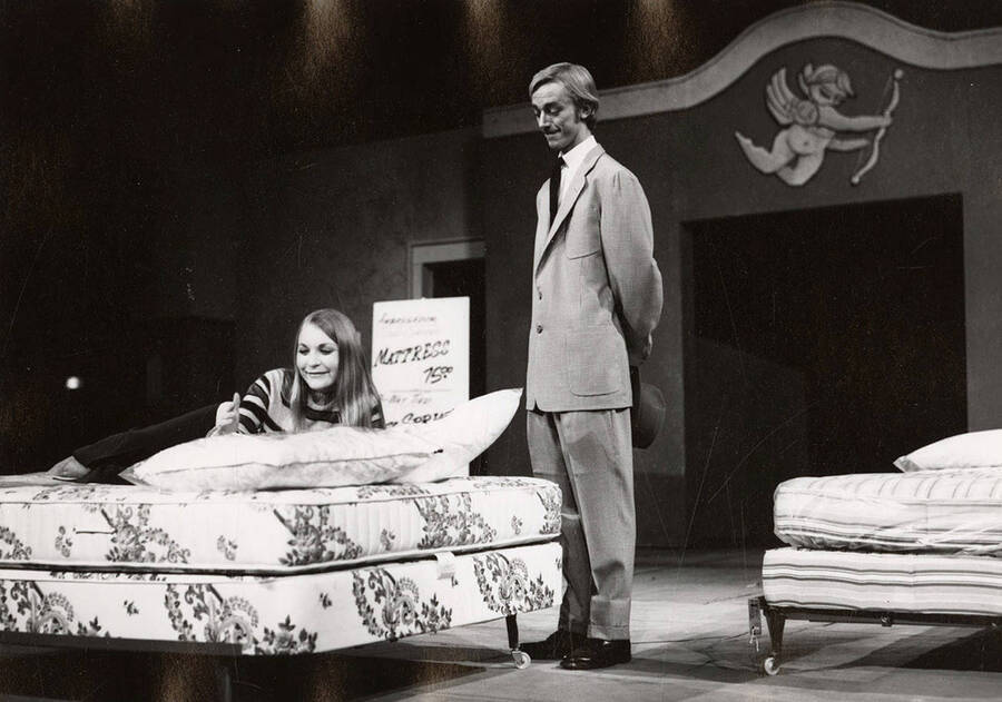A women lays on a mattress while a man stands by looking on during the drama production of 'You Know I Can't Hear You When the Water's Running.'