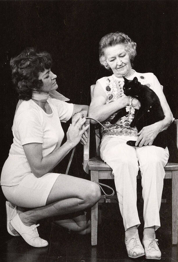 Two women sit on stage holding a cat during a drama production.
