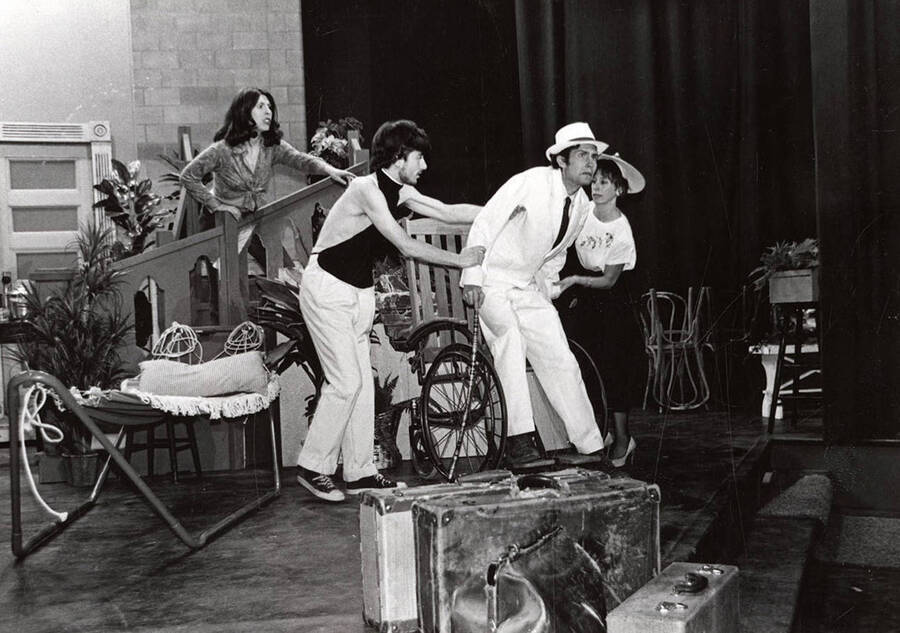 A man helps another man out of his chair, while two women look on during a drama production.