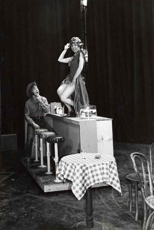 A woman stands on a counter, while a seated man looks on during a drama production.