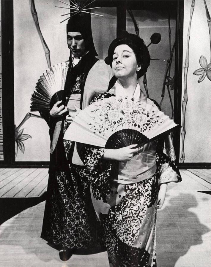 A man and woman stand onstage holding fans during a drama production.