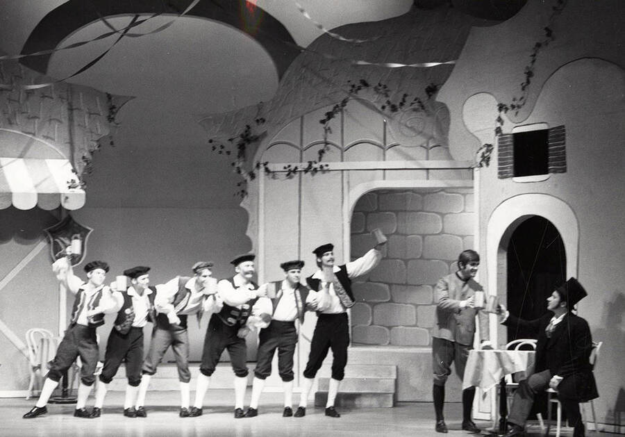 Men stand onstage and raise their mugs in the air during a drama production.