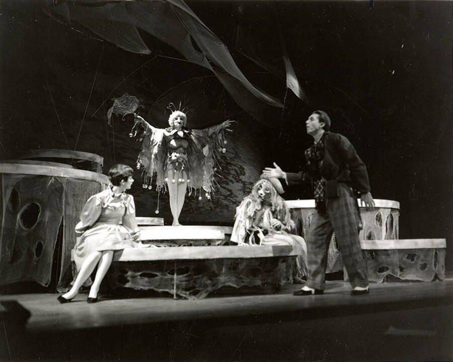 Scene from the University of Idaho drama production of 'The Man in the Moon.' A woman can be seen dressed in costume with her arms spread. Three other actors can be seen conversing with the woman.