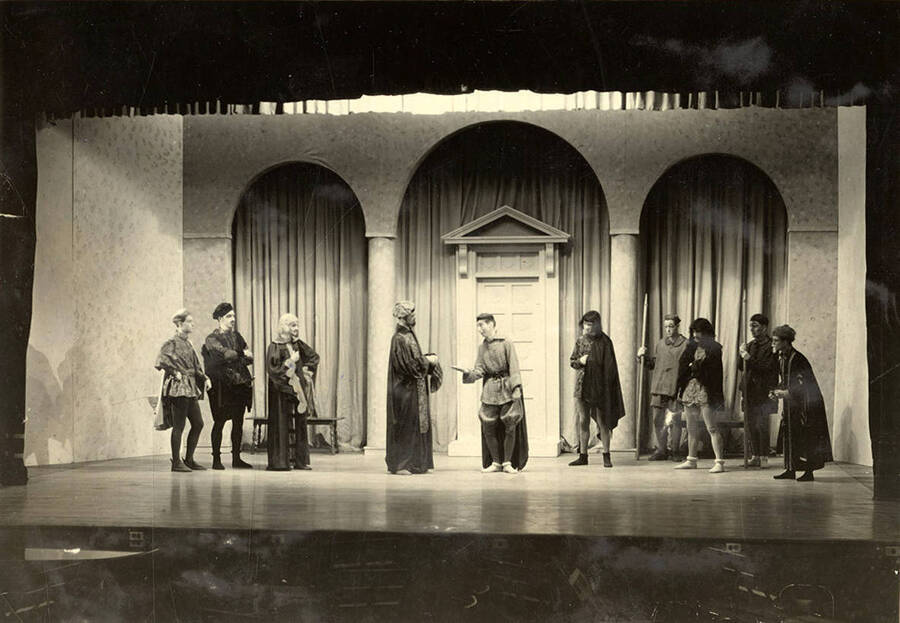 Group scene from the University of Idaho drama production of 'Much Ado About Nothing.' Actors can be seen dressed in costume and conversing on stage. Members of cast identified on mount.