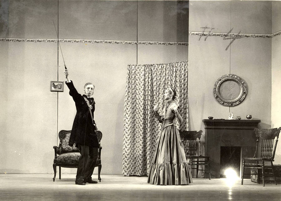 Scene from the University of Idaho drama production of 'Trelawny of the Wells.' A man can be seen holding a prop above his head, while a woman looks on. Members of the cast include Alberta Bergh Utt as Rose Trelawny, Robert Herrick as Tom Wrench, Marion Dresser as Imogen, Clarence Anderson as Colpoys, Jean Ricker as Avonia Bunn and Raphael Gibbs as Sir Gower.