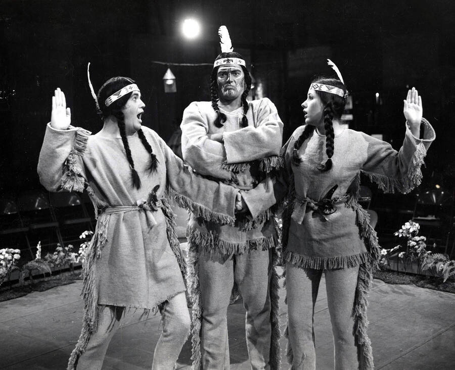 Scene from the University of Idaho drama production of 'Little Mary Sunshine.' Pictured are Charles Walton as Billy Jester, Veryl Alcorn as Yellow Feather, and Jeannie Smith as Nancy. They can all be seen dressed in costume and Varyl and Jeannie are holding up one hand on either side of Charles.