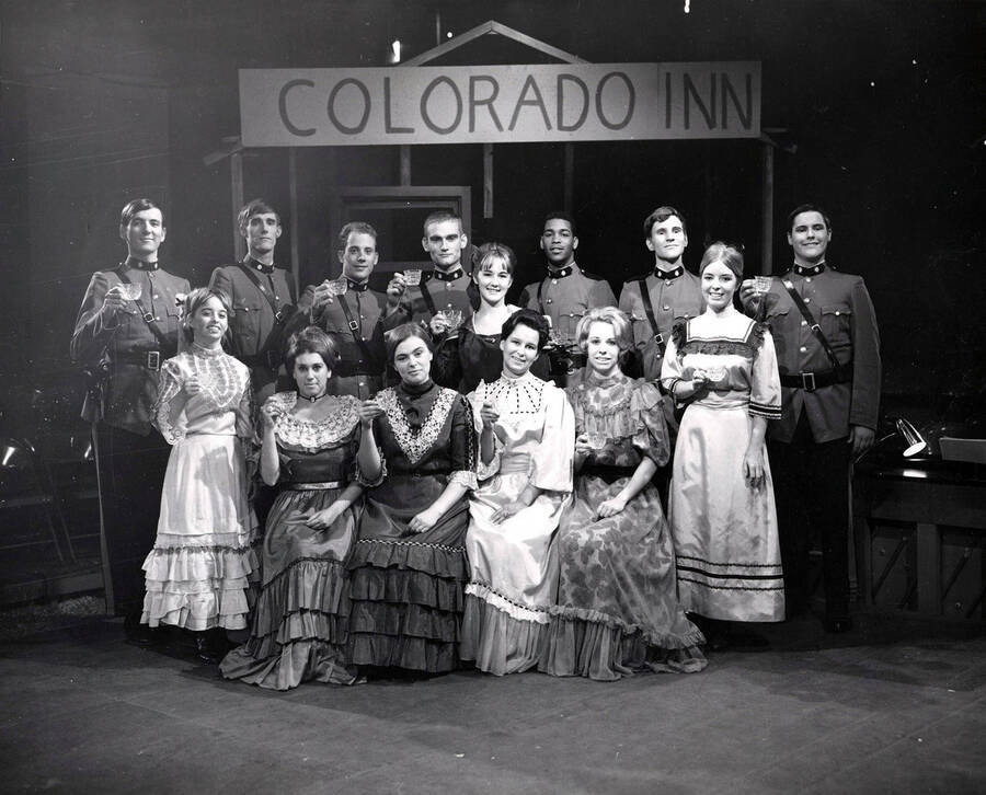 Group photo of the cast from the University of Idaho drama production of 'Little Mary Sunshine.' Women are sitting and standing in front, wearing dresses, and men stand in the back, also in costume. The cast can be seen under a sign that says 'Colorado Inn', holding glasses.